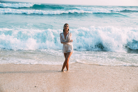 迷人的美丽的女孩在一个时髦的海滩绣礼服站在海滩上, 在高波浪的背景下, 泡沫, 微笑和引诱她的眼睛