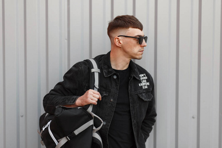 时尚年轻英俊的模特男子黑色眼镜在黑色牛仔夹克与 t恤衫与一个袋子附近的灰色金属墙