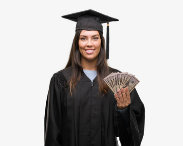 年轻的西班牙裔妇女穿着毕业制服持有美元与幸福的脸站在微笑着自信的微笑显示牙齿
