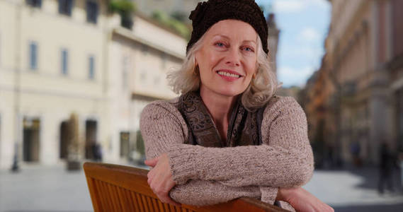 愉快的白人妇女在她的50年代坐在长凳上在城市广场微笑