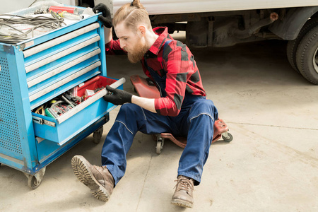 年轻的胡子男子汽车技术员坐在机械爬行者和选择正确的工具, 卡车维修维修店