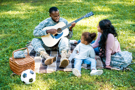 微笑的非洲裔美国人战士在军事制服演奏吉他为家庭在公园