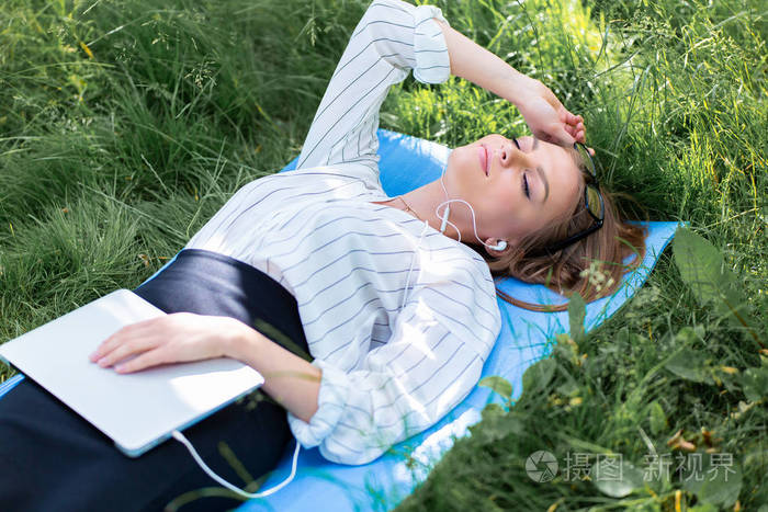 妇女休息在公园躺在草地上的笔记本电脑和听音乐