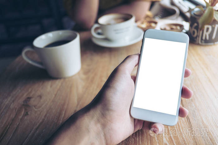 手持白手机的样机图象与咖啡馆里的人一起用空白屏幕和咖啡杯放在木桌