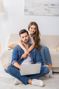 微笑的女人拥抱男朋友, 而他使用膝上型电脑和坐在地板上在家