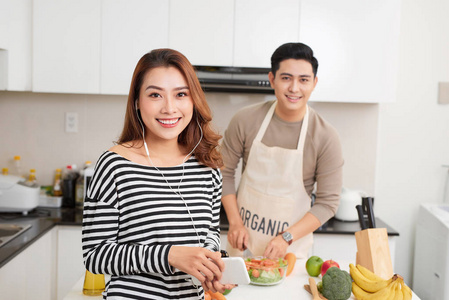 漂亮的年轻夫妇在厨房做饭在家。男子烹饪和妇女使用数码片和微笑