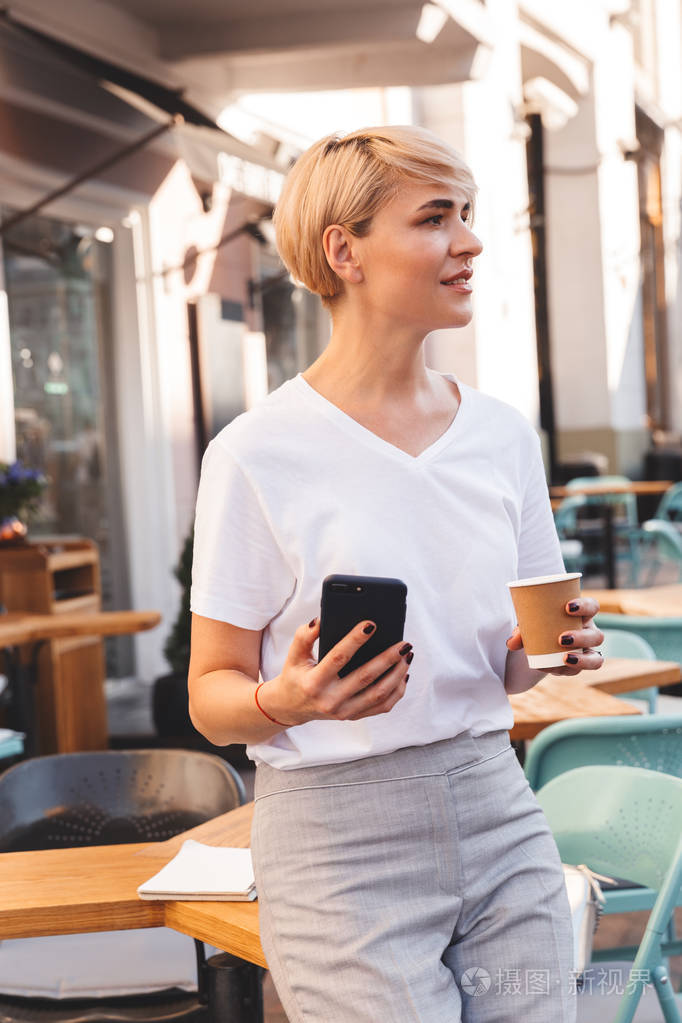穿着白色 t恤衫的漂亮成年女子照片在夏天外面的咖啡馆里坐着, 喝着