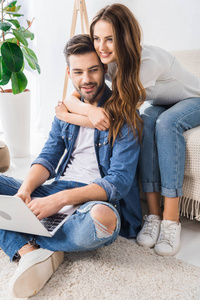 年轻的微笑的女人拥抱男朋友, 而他使用笔记本电脑坐在地板上在家