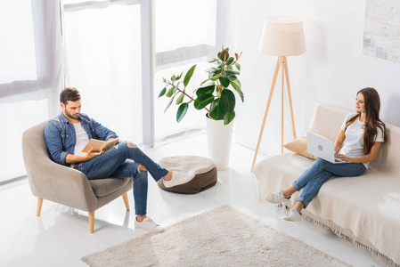 妇女的高角度视图使用膝上型电脑在沙发上, 而她的男朋友在家里的扶手椅阅读书