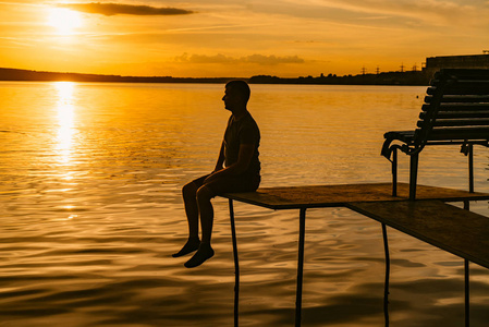 在夏季日落时分, 成年男子坐在砖石上, 在河的背景上放置长凳。美景