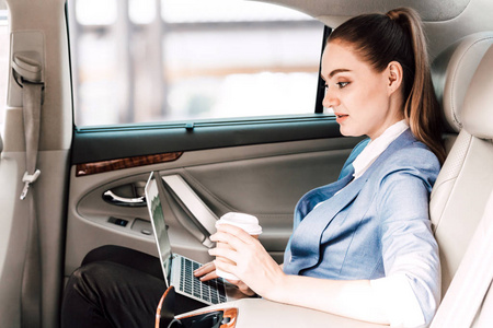 女实业家与笔记本电脑一起工作, 在豪华轿车后座捧杯咖啡