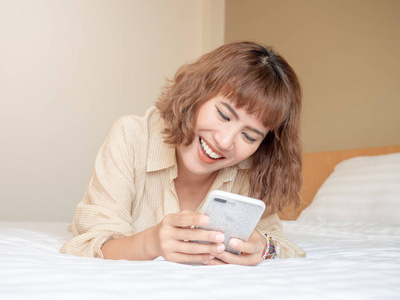 美丽的亚洲女人短发笑躺在白色的床上使用手机