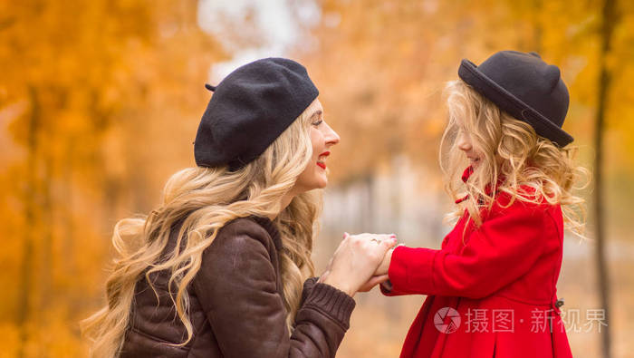 拥抱一个小女儿在明亮的红色大衣和黑色的帽子在金色的秋季花园的背景