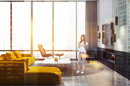 妇女在时尚的客厅内部与白色的墙壁, 木地板, 黄色沙发, 两个黑色扶手椅和电视机挂在壁炉上方。色调图像双曝光