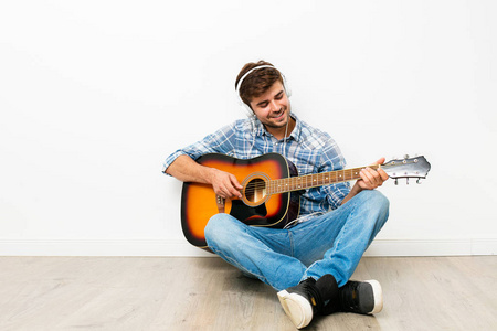 男孩的爱好。男子用耳机在他的头上玩吉他, 坐在地板上, 享受他的业余时间在自己的房子, 做他喜欢的, 在白色的背景