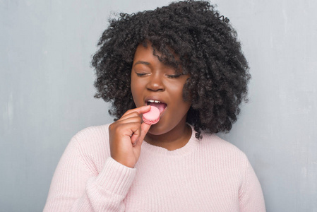 年轻的非洲裔美国妇女在灰色的垃圾墙上吃粉红色的 macaron 与自信的表情, 聪明的脸认真思考
