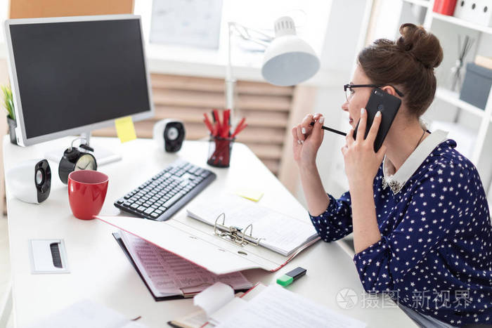 一个戴眼镜的年轻女孩坐在办公室的电脑桌旁, 正在打电话.