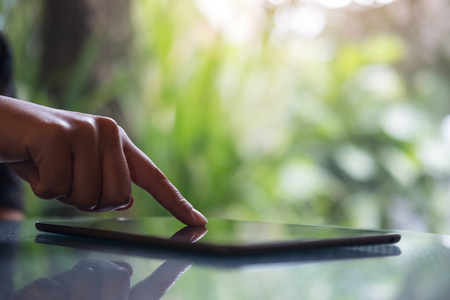一个女人的手指触摸和指向在玻璃桌上的平板电脑与绿色自然背景的咖啡馆模糊背景