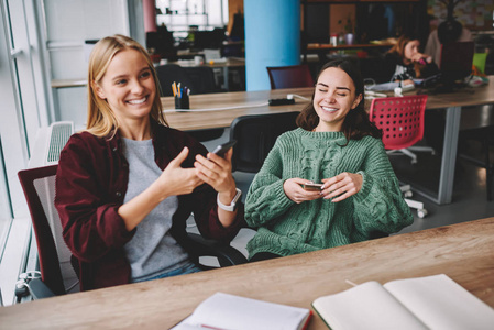 快乐的白种时髦女孩一起在 coworking 空间使用手机和蓝牙连接共享多媒体, 快乐开朗的女性朋友笑在照片上 cellular