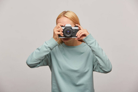女孩覆盖她的脸与相机做一个摄影会议。摄影师拍照, 工作在工作室, 室内摄影, 身着蓝色运动衫, 灰色背景