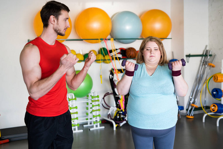 健身房肥胖健身教练在健身房减肥训练中用哑铃做运动的紧张肥胖妇女的