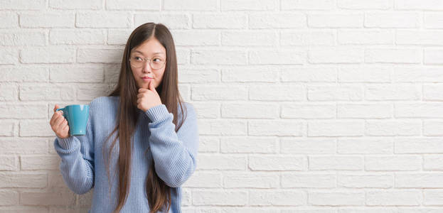 年轻的中国女人喝着一杯咖啡在白砖墙上严肃的脸上想问题, 很迷茫的想法