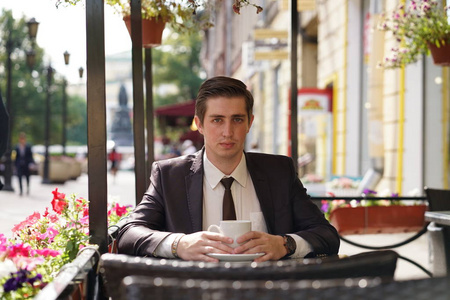 一个穿着黑色西装, 白色衬衫和领带的年轻人坐在一张桌子上的城市街咖啡馆里, 喝着一杯咖啡。