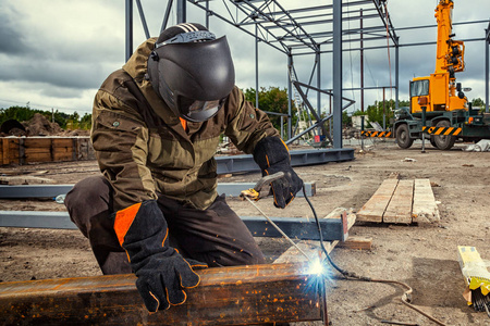 一个年轻男子焊工在棕色制服, 焊接面具和焊工皮革, 焊接金属与电弧焊机在建筑工地上, 蓝色火花飞向两侧