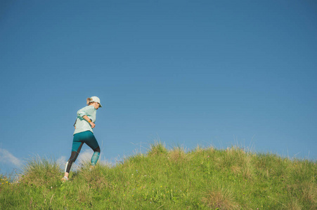 有吸引力的运动女孩在帽子和耳机慢跑通过草地上风景如画的地方, 在山上的蓝天。户外锻炼