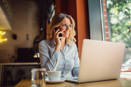 戴眼镜的年轻商界女性的肖像坐在她的笔记本电脑前的咖啡馆, 并谈论手机