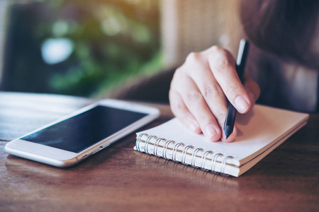 在咖啡桌上用白色手机在笔记本上手持铅笔和写字的妇女特写图像