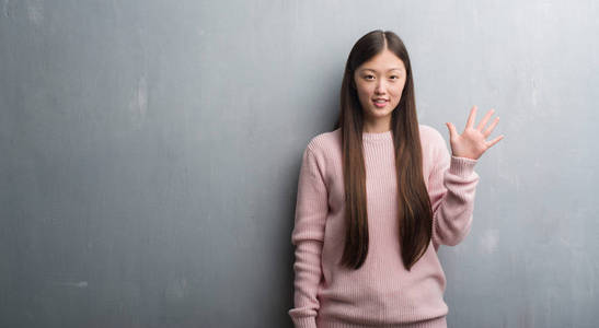 年轻的中国妇女在灰色墙壁显示和指向用手指五, 而微笑自信和愉快