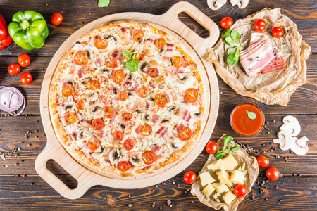 大比萨饼与培根, 蘑菇和西红柿在一个圆形的切菜板上的一个黑暗的木制背景。食品配料