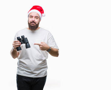年轻的高加索嬉皮士男子戴着圣诞帽看虽然双筒望远镜在孤立的背景非常高兴用手和手指指着