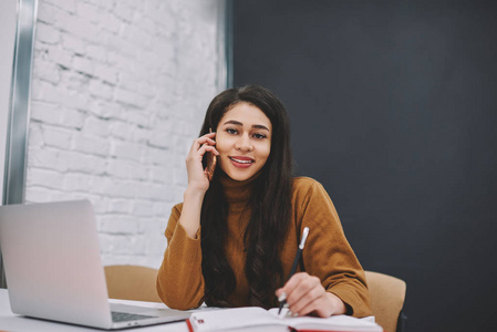 欢快的女学生的肖像坐在 coworking 空间与膝上型计算机, 积极的年轻妇女在手机交谈期间注意到工作中的信息