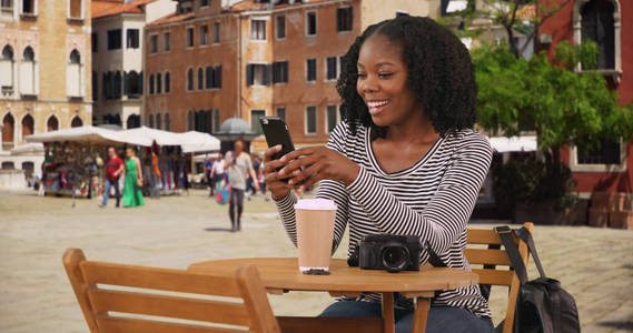 可爱的黑人妇女坐在威尼斯城市街道的咖啡馆桌旁, 在电话里发短信