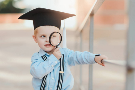 戴眼镜和学术帽的小男孩站在一所学校的砖墙上。持有放大镜并研究环境