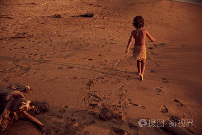 卷发的无忌男孩手里拿着棍子走在海滩上.从后面开枪