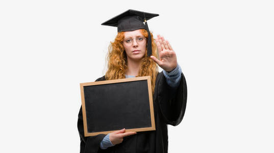 年轻红发学生女子穿着毕业制服拿着黑板张开手做停止标志以严肃自信的表情, 防守姿态