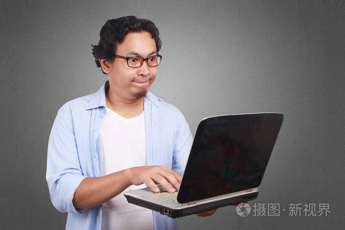 年轻的亚裔男子穿着白色和蓝色的衬衫与愤怒的表情看着 lptop。关闭正文肖像