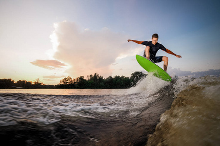 有吸引力的男性 wakeboarder 跳在绿色板上的摩托艇高浪在湖边的背景下