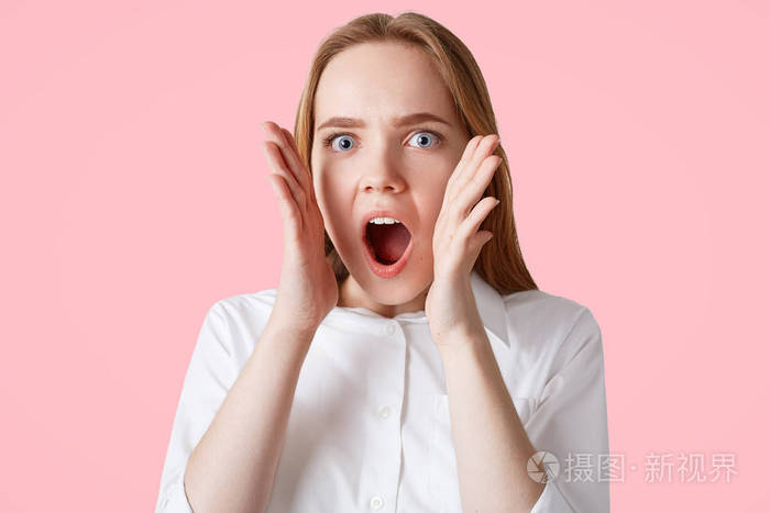 惊奇的蓝眼睛的欧洲女性惊呼以惊恐的表示, 听见假新闻, 反对粉红色