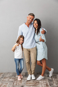 一个幸福的家庭的肖像父亲, 母亲, 小女儿拥抱, 而站在一起, 并显示大拇指上孤立的灰色背景
