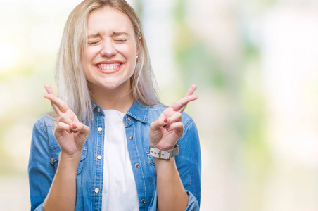 年轻的金发女人在孤立的背景下微笑着交叉手指, 希望和眼睛闭上。运气和迷信概念