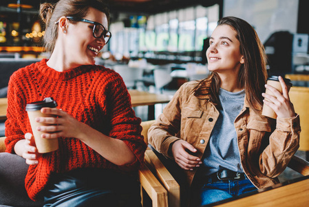 两个快乐的女性人享受现场交流和早晨外卖咖啡在大学食堂, 时髦的微笑