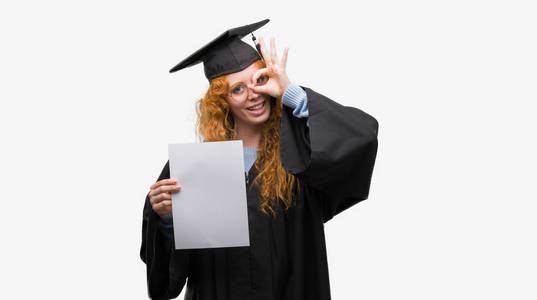 身穿毕业制服的年轻红发女子带着快乐的脸微笑着做 ok 手势用手在眼睛看通过手指
