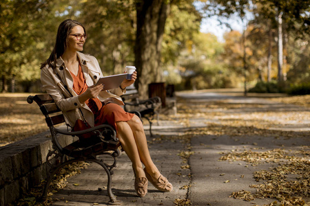 秋季天气, 妇女喝咖啡和坐在公园长椅上, 使用平板电脑和检查社交媒体