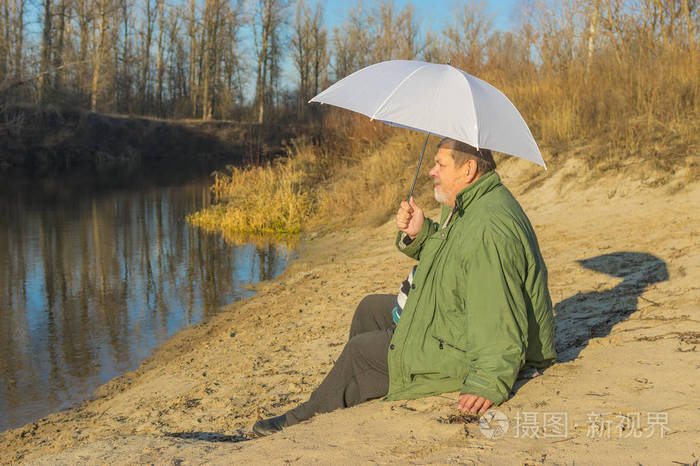 在阳光明媚的秋日 老人带着白色的伞坐在河边的海滩上照片-正版商用
