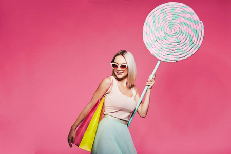 美丽的年轻女子戴着墨镜持有巨大的甜棒糖和五颜六色的购物袋站在粉红色的背景, 面部表情