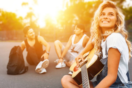 一群朋友在日落的时候在滑冰场的沥青上嬉戏, 特写美丽的微笑的女孩在一群朋友的背景下, 弹吉他, 好夏天的心情